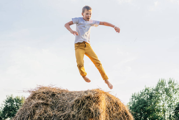 Portrait of barefoot boy in white short and yellow trousers jumping on haystack in field. Outdoor activity. Side view - Foto, Imagen
