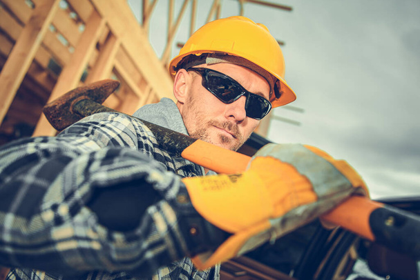 Kavkazský profesionální stavební dělník ve věku 40 let s velkým kladivem na rameni. Nošení žluté tvrdé klobouky a ochrana očí Sluneční brýle. - Fotografie, Obrázek
