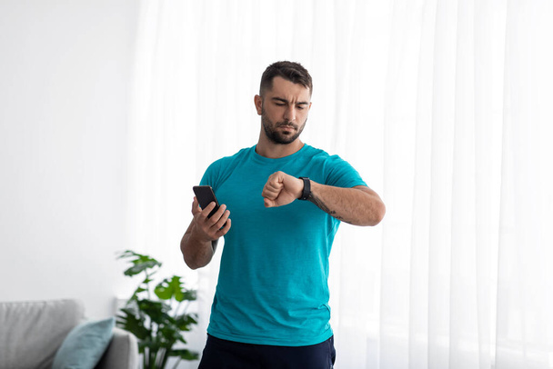 Millenniumi izom európai férfi ellenőrzi pulzus fitness tracker és telefon a szobában belső ablak háttér - Fotó, kép