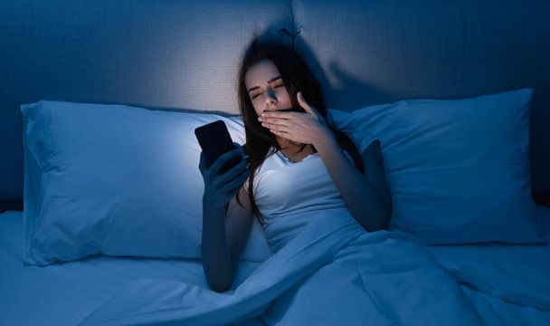 Sleepy female using cellphone in bed at night - Foto, imagen