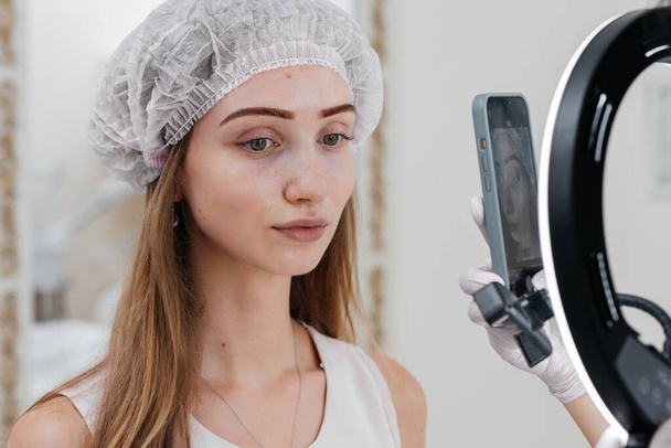 The cosmetologist photographs the patient after the procedure for filling the nasolacrimal furrow and mesotherapy around the eyes for a young beautiful girl. Modern cosmetology. - Photo, Image