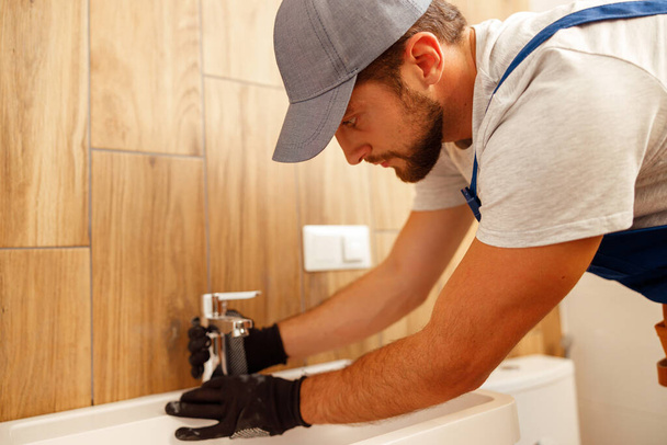 Serieuze loodgieter, mannelijke werknemer in uniform installeren kraan of badkamer kraan - Foto, afbeelding