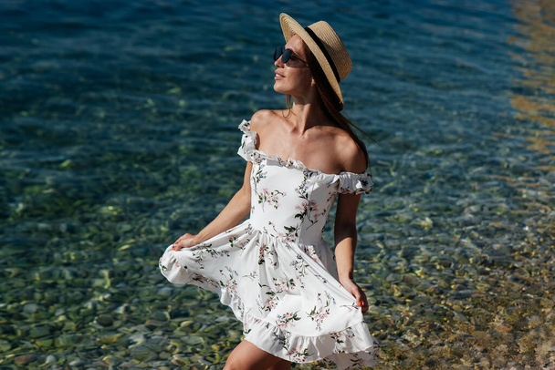 A beautiful young woman in a hat, glasses and a light dress is walking along the ocean shore against the background of huge rocks on a sunny day. Tourism and tourist trips. - Foto, Bild