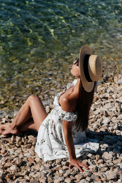 A beautiful young woman in a hat, glasses and a light dress is sitting with her back to the ocean against the background of huge rocks on a sunny day. Tourism and tourist trips. - Photo, Image