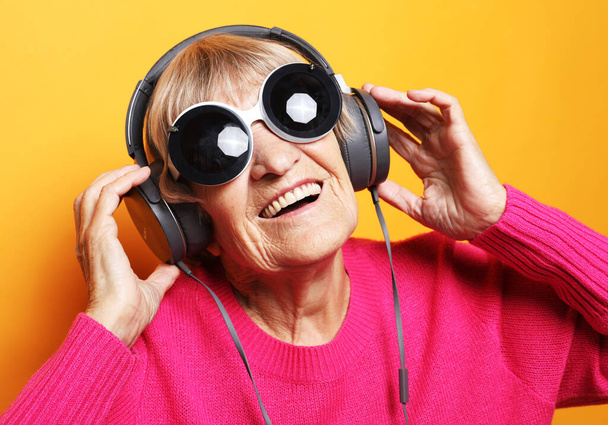 Lifestyle, tehnology and people concept: Funny old woman wearing pink sweater and big sunglasses listening music with headphones over yellow background - Фото, изображение