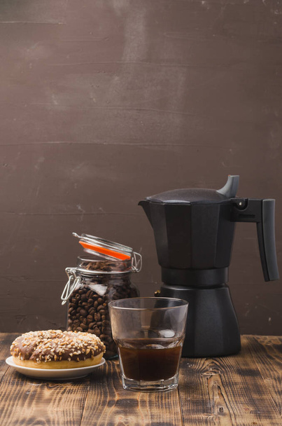Cup of cofee with cofee beans and black maker. Selective focus. Cup of black coffee in a cup, donut with nuts and grains in a glass jar with a coffee maker on a brown background. - Foto, Imagem