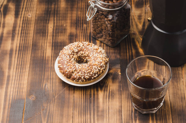 Cup of cofee with cofee beans and black maker. Top view. Cup of black coffee in a cup, donut with nuts and grains in a glass jar with a coffee maker on a wooden background. - Photo, image