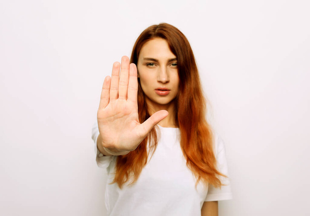 Portrait of a serious young redhair woman standing with outstretched hand showing stop gesture isolated over white background - 写真・画像
