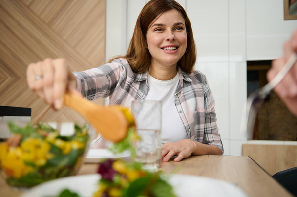 Houkutteleva Kaunis valkoihoinen nainen, kotiäiti, joka tarjoaa terveellistä herkullista vegaanisalaattia valkoisella lautasella ja hymyilee pöydässä kotonaan. - Valokuva, kuva