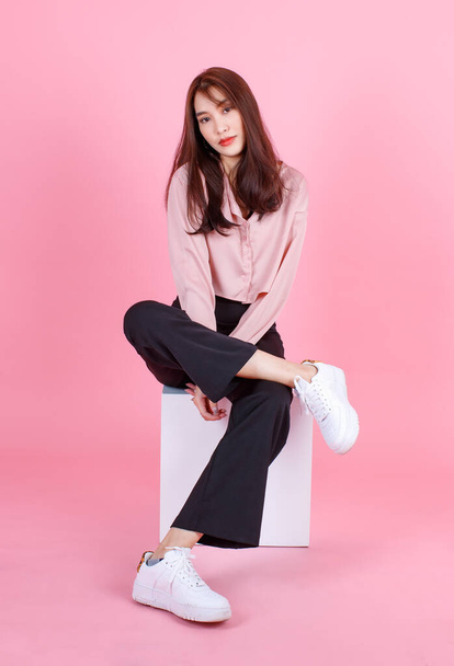 Portrait studio shot of Asian young beautiful hipster female teen model in casual street wears sneakers and sunglasses posing smiling look at camera sit on white square box stool on pink background. - Photo, image
