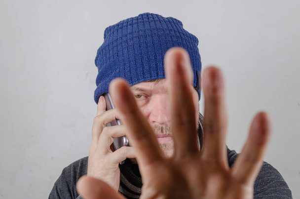Un homme se couvre le visage avec sa paume en parlant sur son téléphone portable. Une personne portant un chapeau bleu contre un mur de couleur claire. À l'intérieur. Concentration sélective. - Photo, image