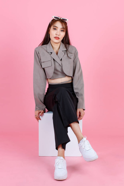Portrait-Studioaufnahme von asiatischen jungen urbanen trendigen weiblichen Hipster Teenager-Mode-Model in lässigen Crop Top Street trägt Jacke mit Sonnenbrille auf dem Kopf sitzen auf weißen quadratischen Kastenhocker auf rosa Hintergrund - Foto, Bild