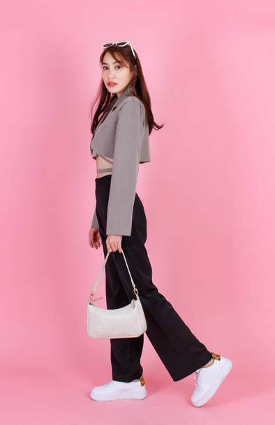 Portrait studio shot of Asian trendy fashionable female hipster teen model in casual crop top street wears jacket sunglasses sneakers holding handbag purse walking look at camera on pink background. - Photo, Image