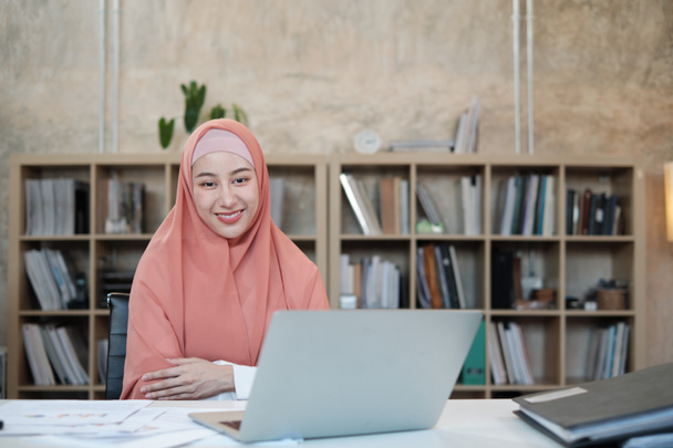 İslami kadın tesettürlü, tesettürlü ve küçük bir ofis iş yerinde kameraya bakan, beyaz masadaki dizüstü bilgisayarla çalışan, arkasında kitaplık olan güzel bir iş adamının portresi.. - Fotoğraf, Görsel