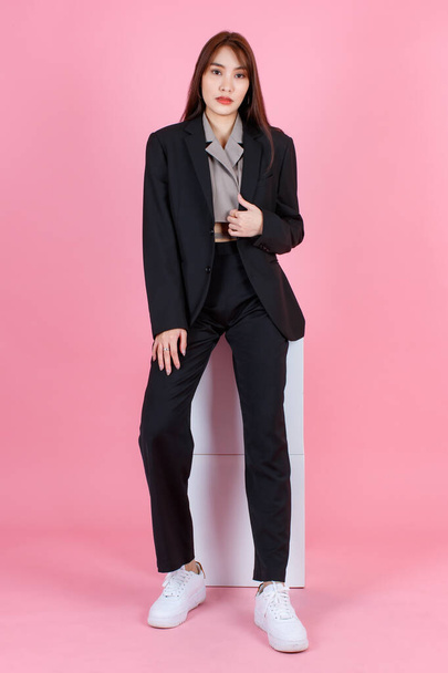 Portrait studio shot of Asian beautiful urban trendy modern fashionable long hair female hipster teenager model in casual black suit with crop top shirt sitting look at camera on pink background. - Photo, Image