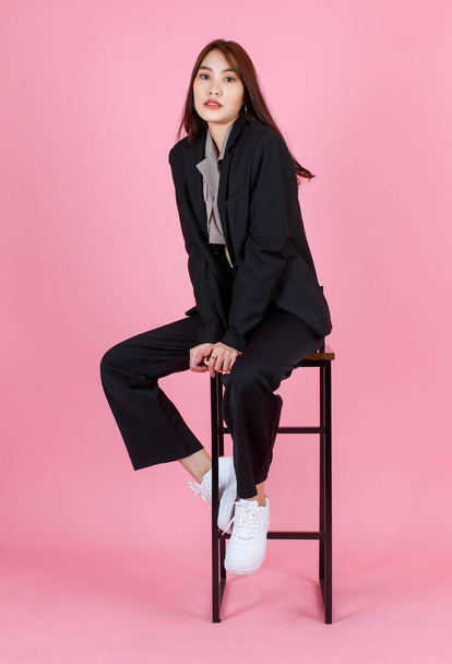 Portret studio shot van Aziatische mooie urban trendy moderne modieuze lange haar vrouwelijke hipster tiener model in casual zwart pak met gewas top shirt zit te kijken naar de camera op roze achtergrond. - Foto, afbeelding