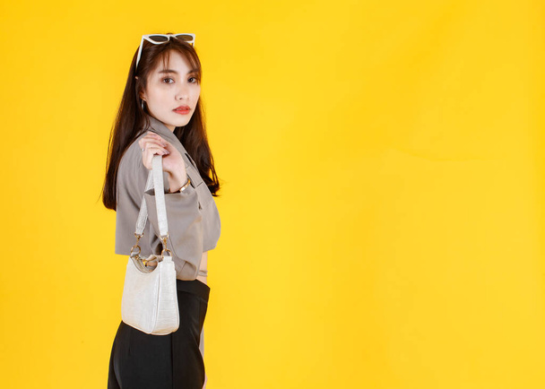 Portrait studio shot of Asian trendy fashionable female hipster teen model in casual crop top street wears jacket sunglasses sneakers holding handbag purse look at camera on yellow background - Photo, Image