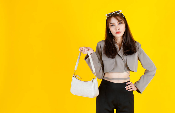 Portrait studio shot of Asian trendy fashionable female hipster teen model in casual crop top street wears jacket sunglasses sneakers holding handbag purse look at camera on yellow background - Photo, Image