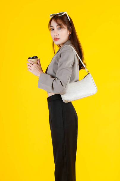 Retrato estudio de toma de moda urbana asiática mujer de negocios en traje de oficina con gafas de sol de pie mirar a la cámara que lleva bolso de cuero monedero y taza de café desechable sobre fondo amarillo. - Foto, Imagen