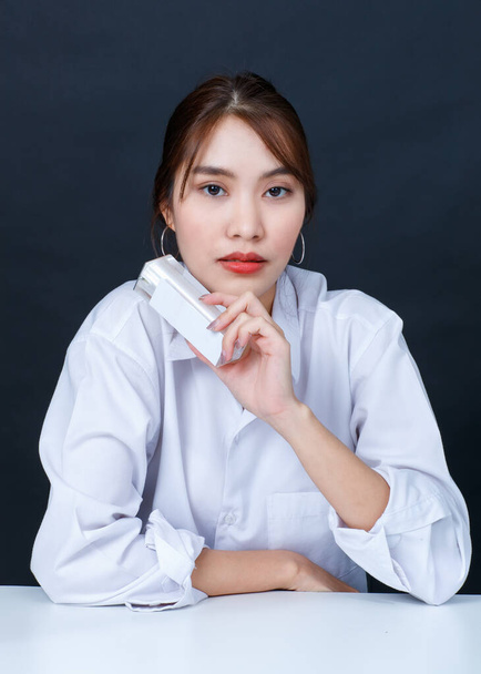 Portré closeup stúdió lövés ázsiai fiatal glamour trendi divatos női modell visel smink alkalmi fehér ing ül mosolygós nézd kamera gazdaság arc krém palack fekete háttér - Fotó, kép