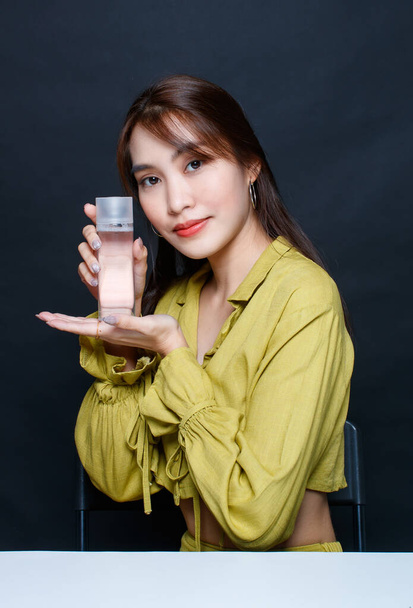 Portrait closeup studio shot Asian glamour trendy fashionable female model wearing makeup in casual outfit look at camera holding no brand mineral water treatment or toner product on black background. - Photo, image
