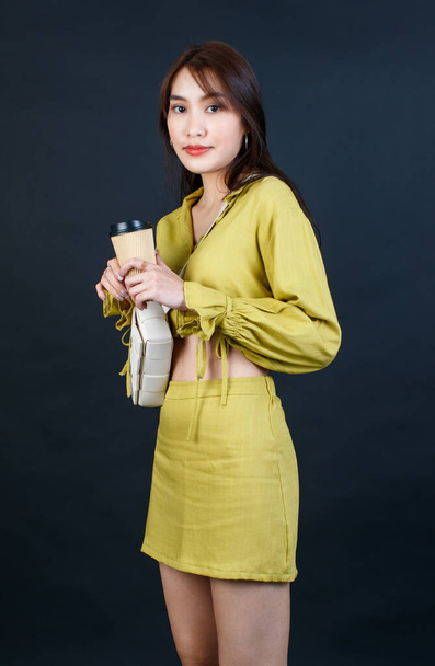 Portrait-Studioaufnahme asiatischer urbaner Mode weibliche Geschäftsfrau in lässigem Crop-Top-Outfit mit Handtasche Handtasche in der Hand, Einweg-Kaffeetasse Blick in die Kamera auf schwarzem Hintergrund. - Foto, Bild