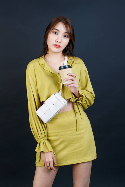 Portrait-Studioaufnahme asiatischer urbaner Mode weibliche Geschäftsfrau in lässigem Crop-Top-Outfit mit Handtasche Handtasche in der Hand, Einweg-Kaffeetasse Blick in die Kamera auf schwarzem Hintergrund. - Foto, Bild