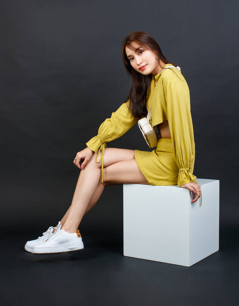 Portrait studio shot of Asian trendy modern fashion female model in casual crop top outfit sitting smiling on white box stool look at camera holding hands pointing fingers sideway on black background. - Photo, Image