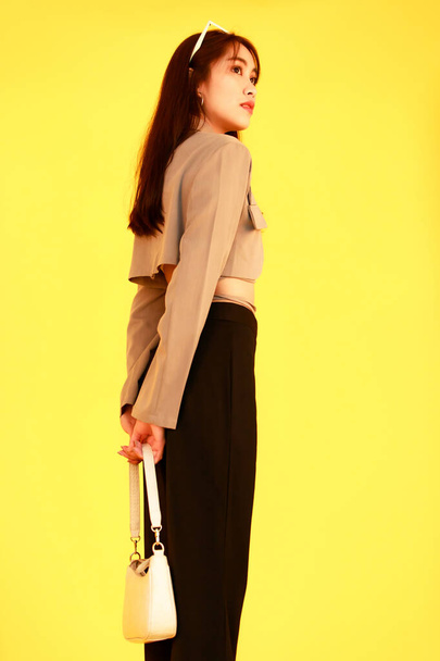 Portrét studio záběr asijské trendy módní žena hipster teen model v ležérní plodina top ulice nosí bundu sluneční brýle tenisky držení kabelka pohled na kameru na žlutém pozadí - Fotografie, Obrázek