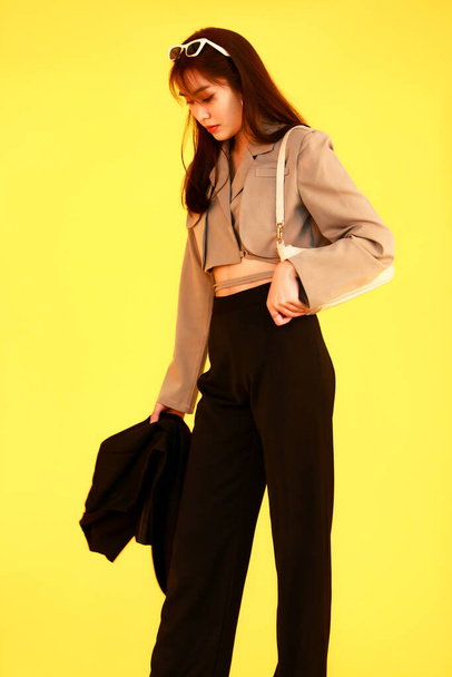 Retrato de estudio de tiro de moda asiática modelo adolescente hipster femenino de moda en la parte superior de las cosechas casual calle lleva chaqueta gafas de sol zapatillas de deporte sosteniendo bolso monedero mirar a la cámara en el fondo amarillo - Foto, Imagen