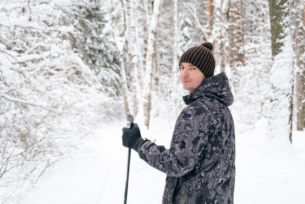 Skier in hat with pompom with ski poles in his hands with his back against the background of a snowy forest. Cross-country skiing in winter forest, outdoor sports, healthy lifestyle. - Foto, imagen