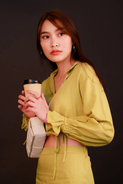 Portret studio schot van Aziatische stedelijke mode vrouwelijke zakenvrouw in casual gewas top outfit dragen handtas portemonnee, met wegwerp koffiekop blik op camera op zwarte achtergrond. - Foto, afbeelding