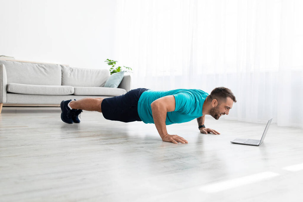 Cheerful millennial muscular european man doing push ups and watching online lesson on computer in living room - Foto, Imagen
