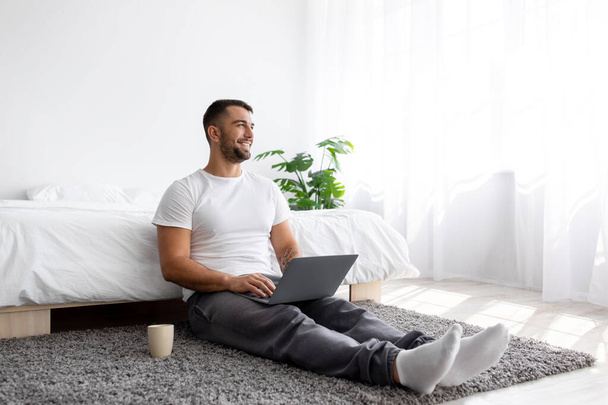 Smiling millennial muscular european male creating idea and working on laptop in bedroom interior - Photo, Image