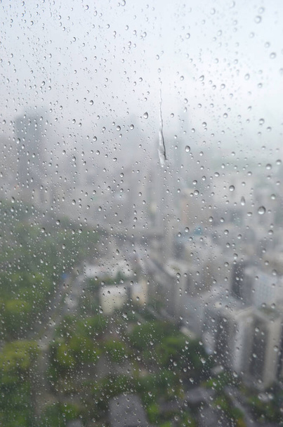 街の建物や木々の緑は、雨の降る窓からしか見えません。雨滴がペインを走っています. - 写真・画像