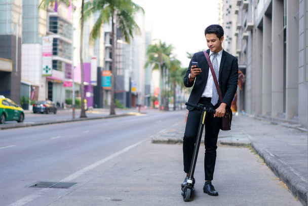 Asiatische Geschäftsmann fährt einen Elektroroller und verwendet sein Handy, um eine Karte in Anwendung zu öffnen, um die Straßen der Stadt zu überprüfen, um morgens zur Arbeit zu fahren - Foto, Bild