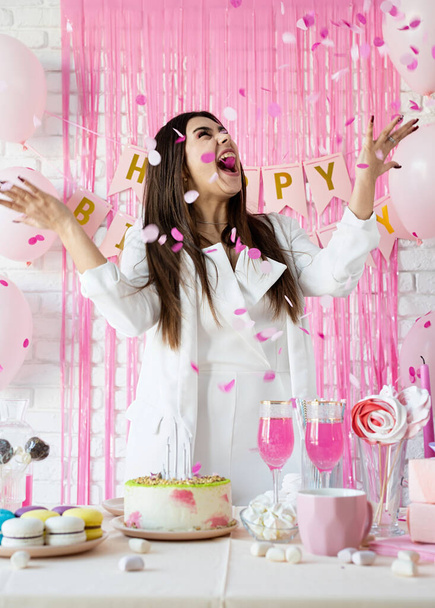 День рождения. Столы на день рождения. Привлекательная брюнетка в белой одежде для вечеринок готовит праздничный стол с тортами, пирожными, макаронами и другими сладостями, бросает розовые конфетти - Фото, изображение