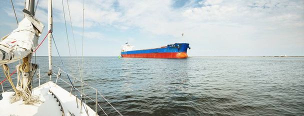 Duży niebieski masowiec (statek towarowy) pływający po Morzu Bałtyckim do portu w Rydze, Łotwa. Widok z jachtu. Dramatyczne zachmurzenie. Komunikacja globalna, logistyka, temat przemysłu - Zdjęcie, obraz