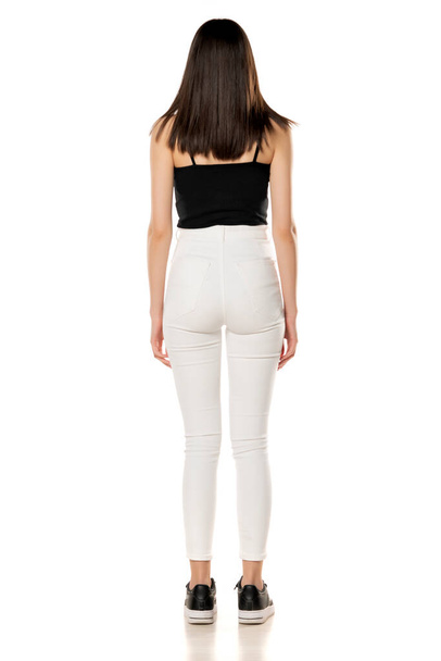 achteraanzicht van een tienermeisje in witte jeans en een zwart shirt staat op een witte achtergrond - Foto, afbeelding