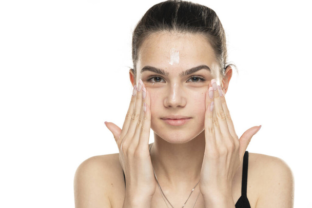 lachende tiener meisje van toepassing cosmetische product op haar gezicht op witte achtergrond - Foto, afbeelding