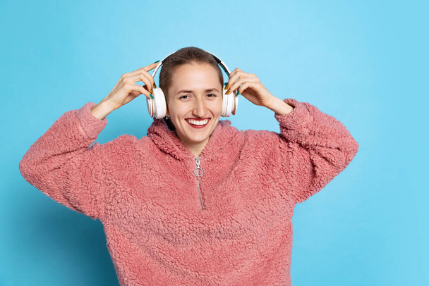 Happy pretty girl, student with short hair wearing pink hoodie in headphones isolated on blue background. Concept of emotions, music - Zdjęcie, obraz