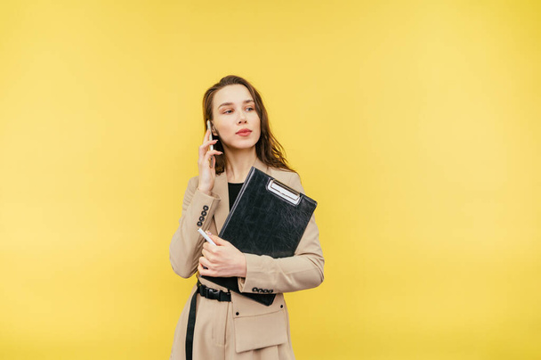 Beautiful business woman in a jacket with a tablet in her hands stands on a yellow background and talks on the phone with a serious face and looks away. - Photo, Image