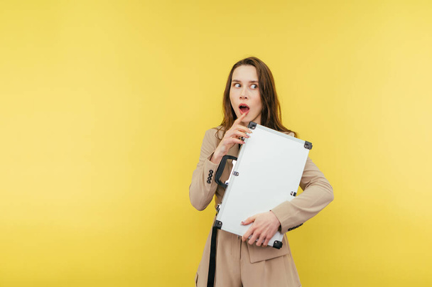 Shocked business woman in a suit with a metal case in her hands stands on a yellow background and looks away with a surprised face. - Foto, Imagen