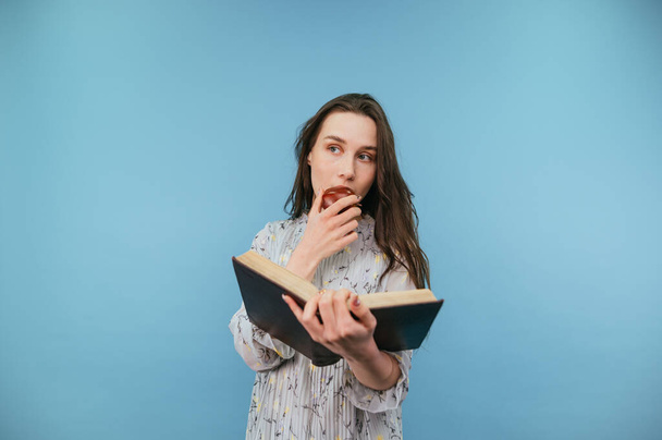 Beautiful woman in a dress with dark hair reads a book and eats an apple on a blue background, looking away with a serious face. - Foto, imagen
