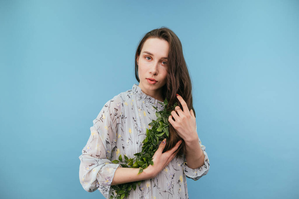 Bella donna bruna in un vestito con una pianta aderente tra le mani isolata su uno sfondo blu, guardando la fotocamera con un viso serio. - Foto, immagini