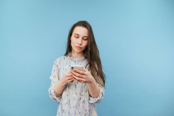 Beautiful dreamy woman in a dress enjoys a smartphone with a serious face on a blue background. Isolated. - Photo, image