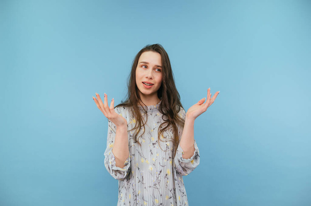 Portrait of a puzzled brunette in a dress on a blue background, looks away and spreads his arms away from confusion, hides his eyes - Photo, Image