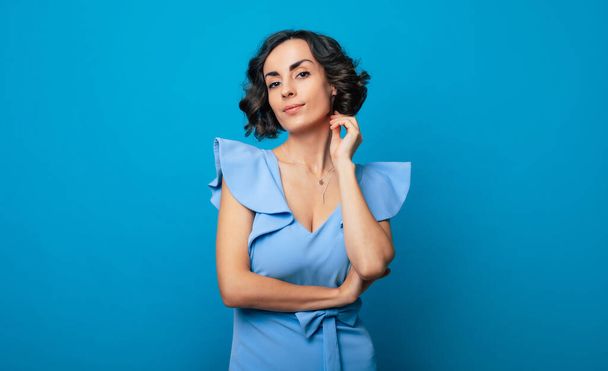 Close-up photo of a charming healthy woman in a blue dress, who is looking in the camera and smiling, while standing and posing isolated on blue background - Foto, afbeelding