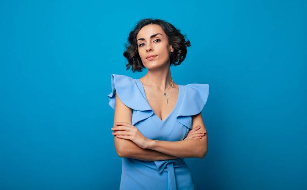 Close-up photo of a charming healthy woman in a blue dress, who is looking in the camera and smiling, while standing and posing isolated on blue background - Photo, image