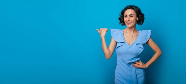 Close-up photo of a happy beautiful young woman in a blue dress, who is pointing with help of her finger and isolated on blue background - Foto, Imagen